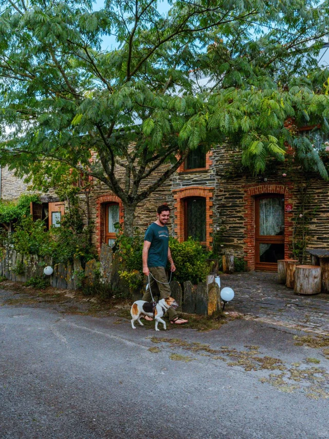 Promeneur et son chien passant devant un gîte en pierre avec terrasse arborée